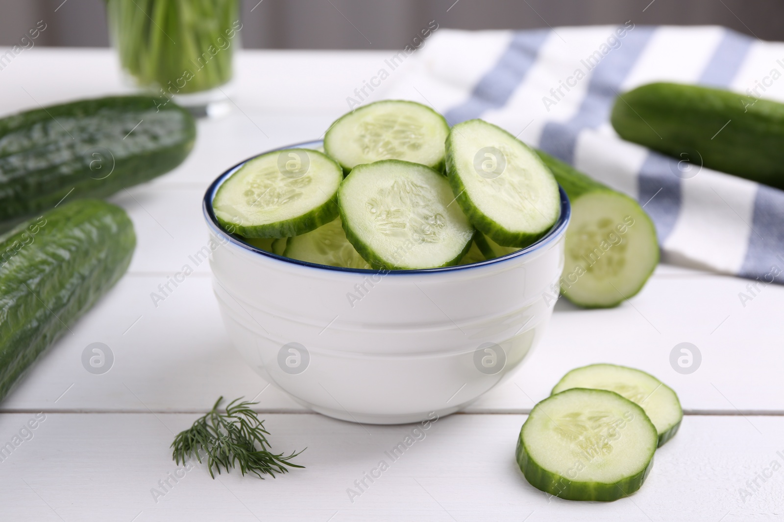 Photo of Cut cucumber in bowl, fresh vegetables and dill on white wooden table, closeup