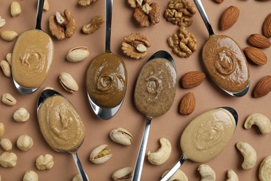 Tasty nut butters in spoons and raw nuts on light brown table, flat lay