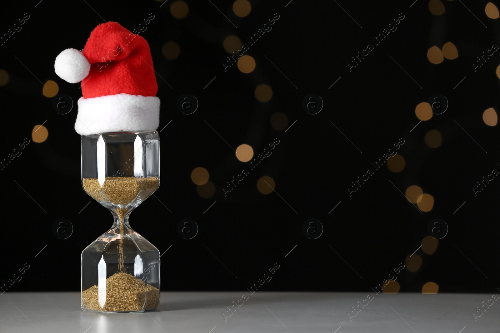 Photo of Hourglass with Santa hat on table against blurred lights, space for text. Christmas countdown