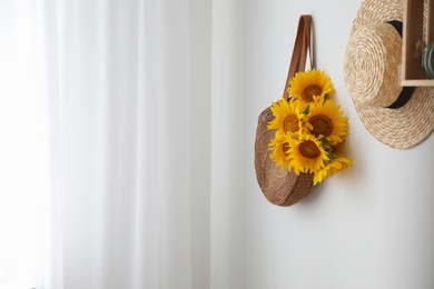 Photo of Knitted bag with bouquet of beautiful sunflowers and straw hat on wall indoors. Space for text