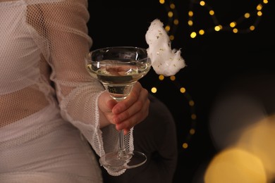 Photo of Woman holding glass of cocktail decorated with tasty cotton candy on black background with blurred lights, closeup. Space for text