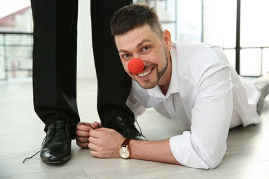 Man with clown nose tying shoe laces of his colleague together in office, closeup. Funny joke