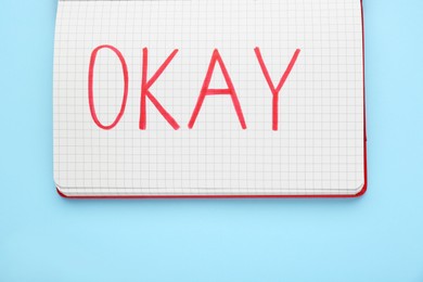 Photo of Notebook with word Okay on light blue background, top view