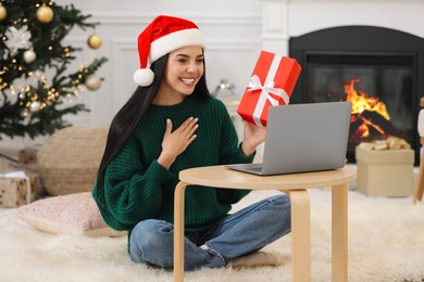 Photo of Celebrating Christmas online with exchanged by mail presents. Smiling woman in Santa hat thanking for gift during video call at home