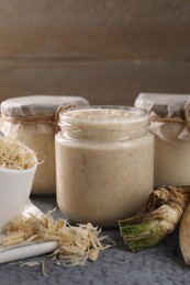 Photo of Spicy horseradish sauce in jars and roots on grey wooden table