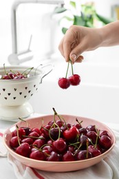 Woman holding fresh ripe cherries over bowl in kitchen, closeup