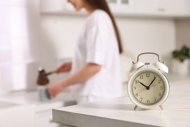 Photo of Alarm clock on white table. Woman pouring coffee from jezve into cup in kitchen, selective focus