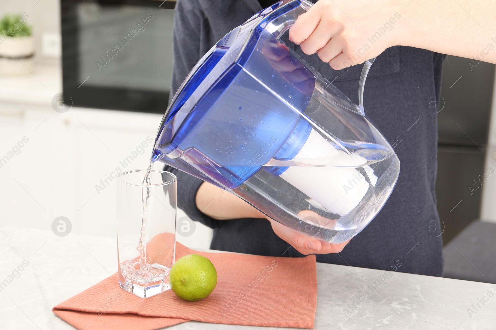 Photo of Woman pouring water from filter jug into glass at light marble table in kitchen, closeup