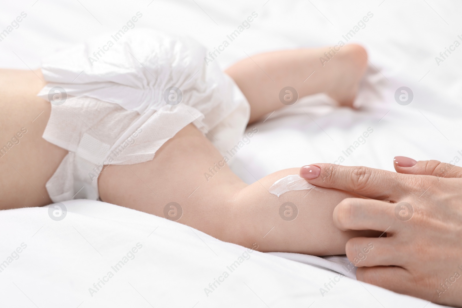 Photo of Woman applying body cream onto baby`s leg on bed, closeup