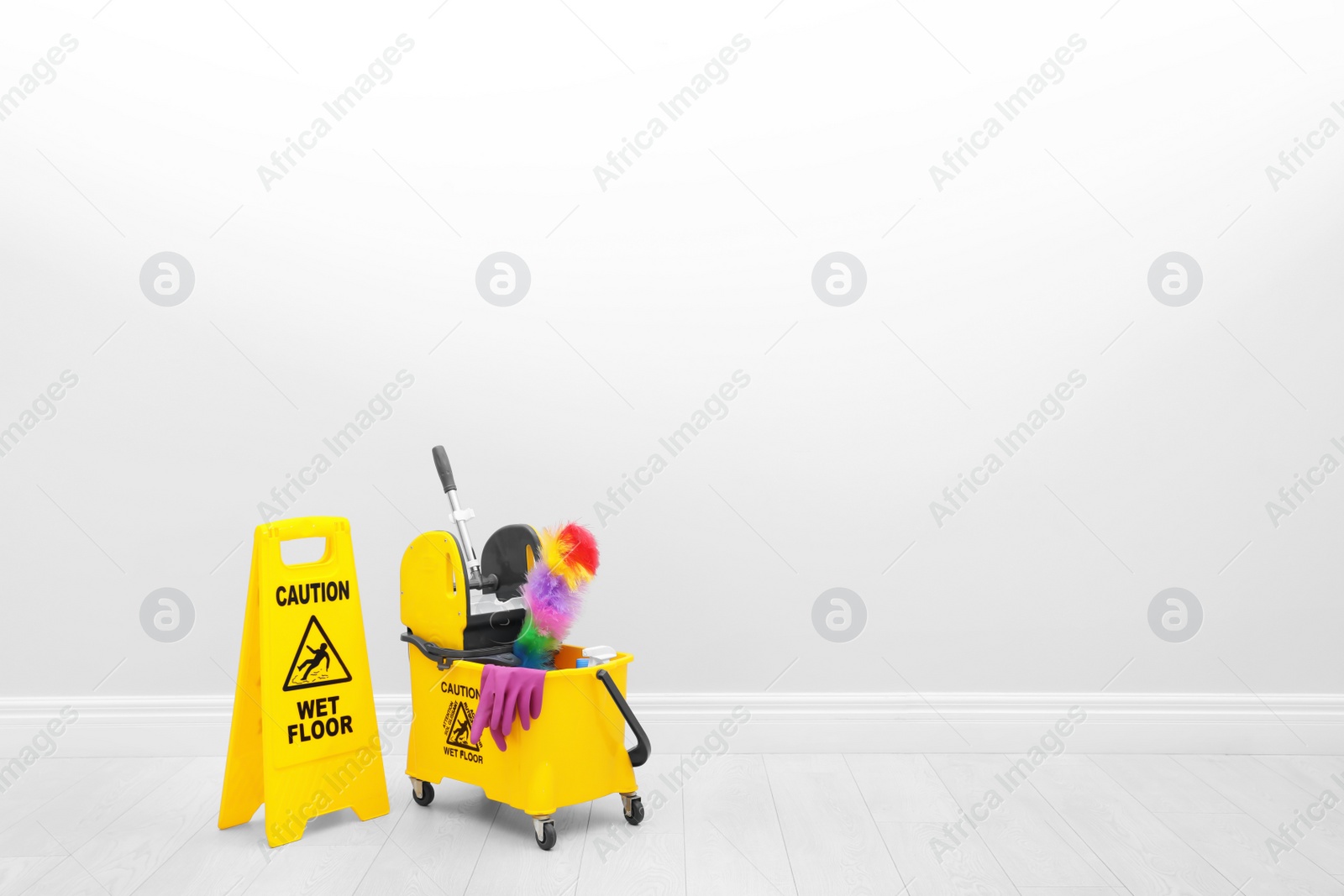 Photo of Safety sign with phrase "CAUTION WET FLOOR" and mop bucket on floor near white wall. Cleaning tools