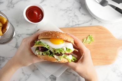 Photo of Woman holding tasty burger with fried egg over table, top view