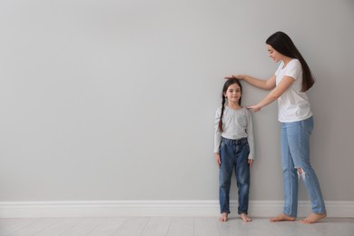 Photo of Mother measuring little girl's height near light grey wall indoors. Space for text