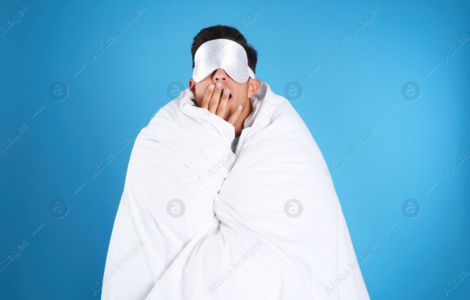 Photo of Man wrapped with blanket in sleeping mask yawning on blue background