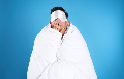 Man wrapped with blanket in sleeping mask yawning on blue background