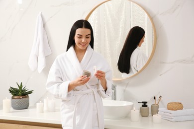 Beautiful young woman with jar of cream near mirror in bathroom