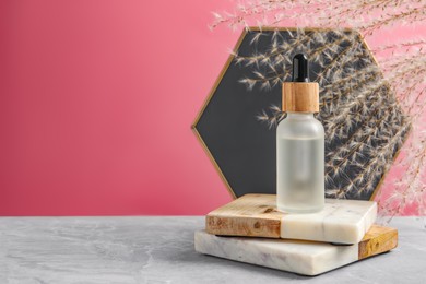 Bottle of face serum with marble boards on grey table against pink background. Space for text