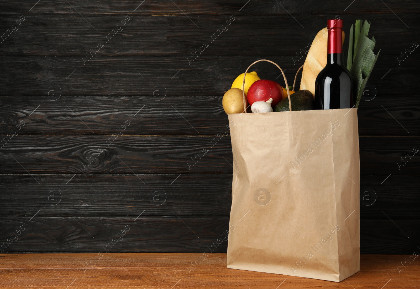 Photo of Paper bag with groceries on wooden table against dark background. Space for text