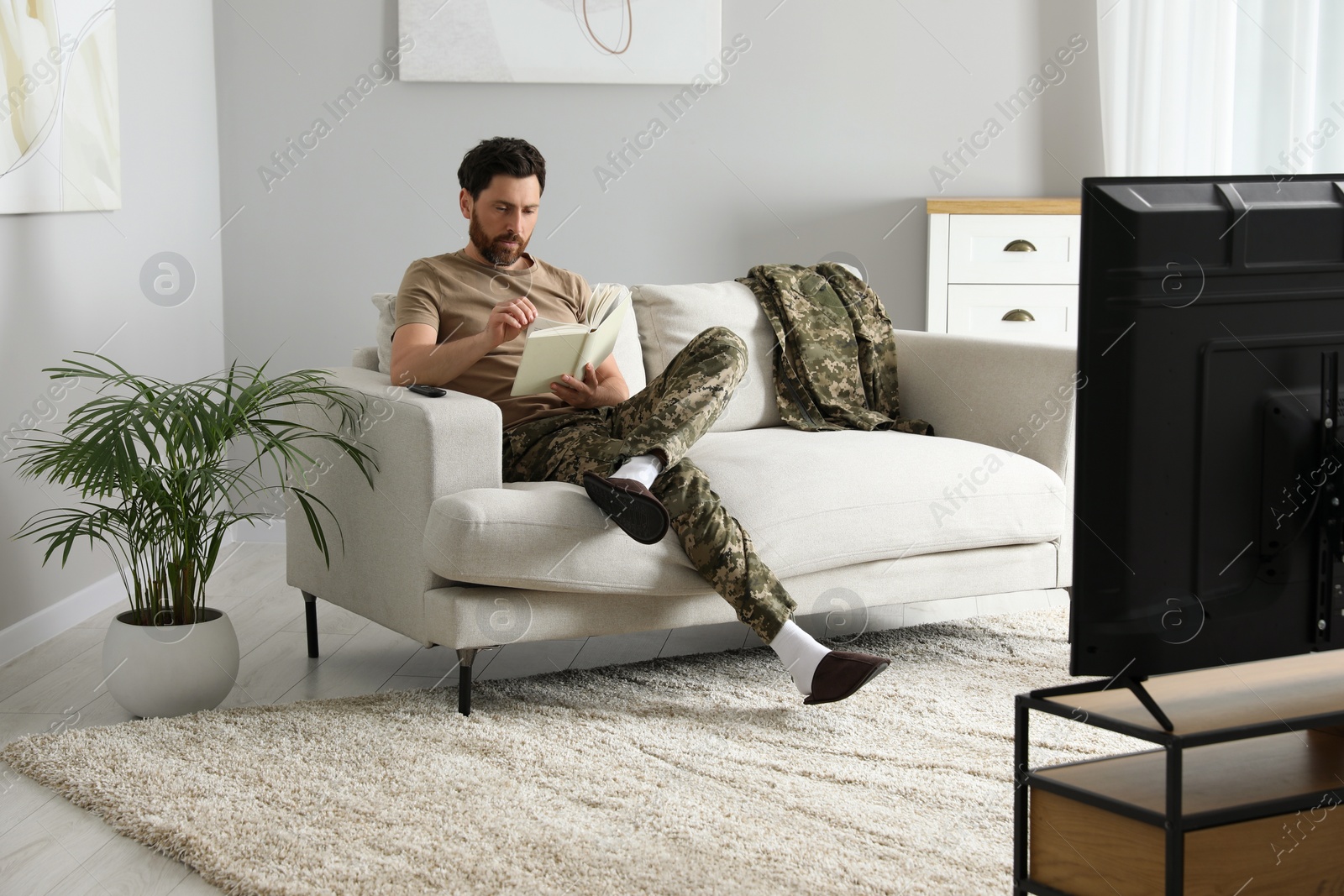 Photo of Soldier reading book on soft sofa in living room. Military service