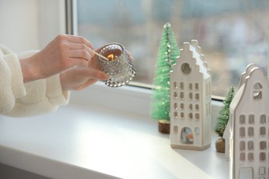 Photo of Woman lightning up candle near windowsill with house shaped lanterns indoors, closeup. Space for text