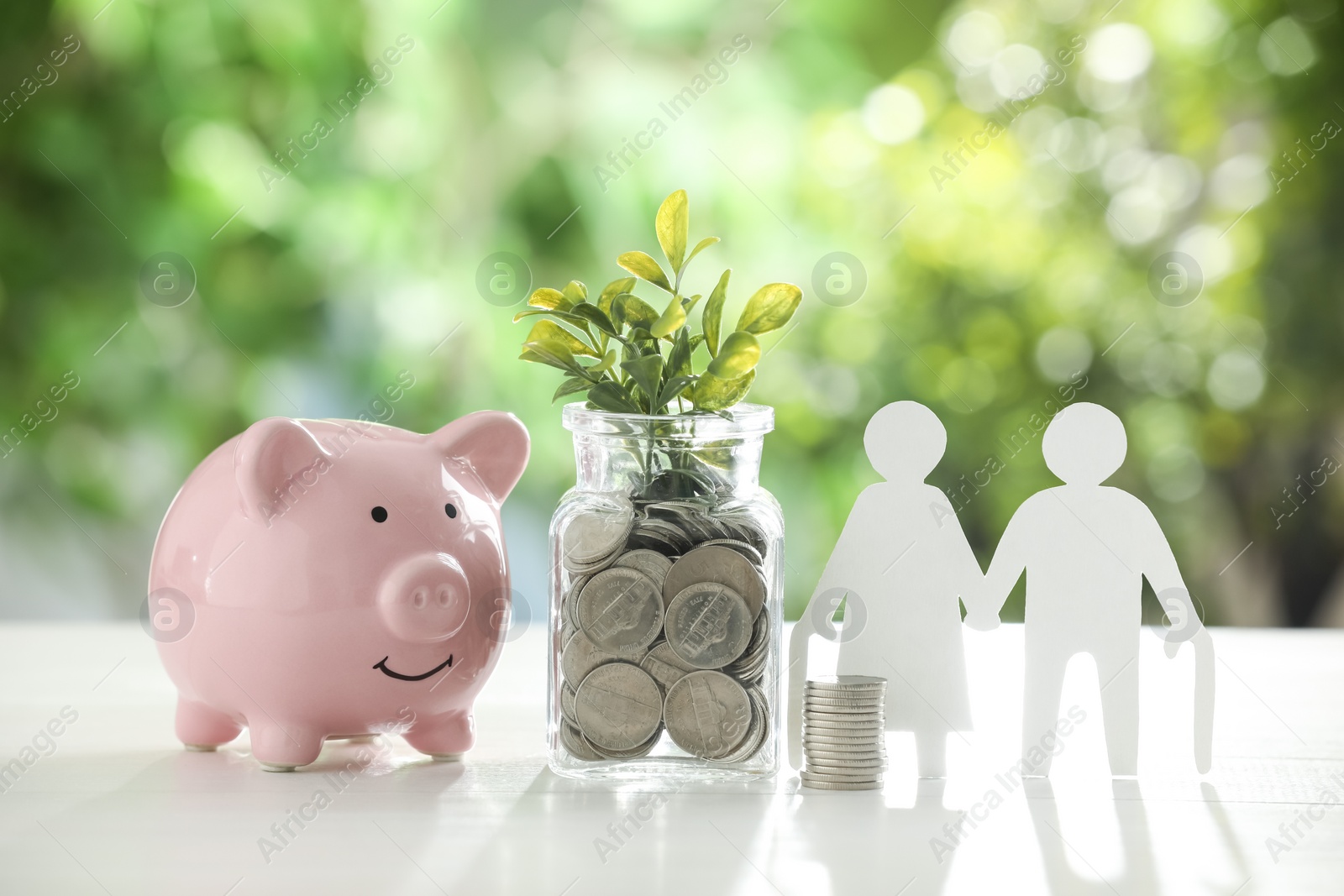 Photo of Pension savings. Figure of senior couple, piggy bank, jar with coins and twigs on white table against blurred green background