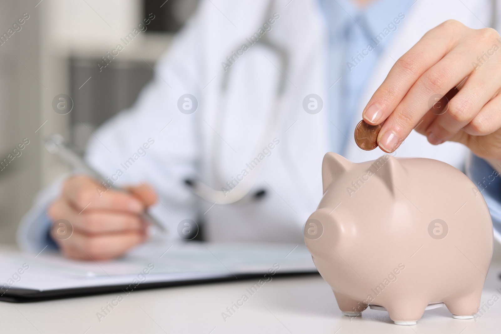 Photo of Doctor putting coin into piggy bank while making notes at white table indoors, closeup