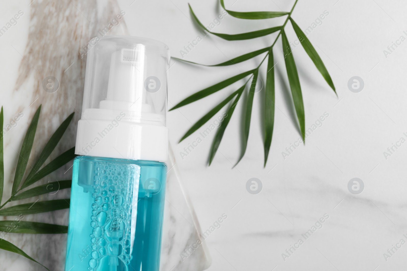 Photo of Bottle of face cleansing product and green leaves on white marble table, flat lay. Space for text