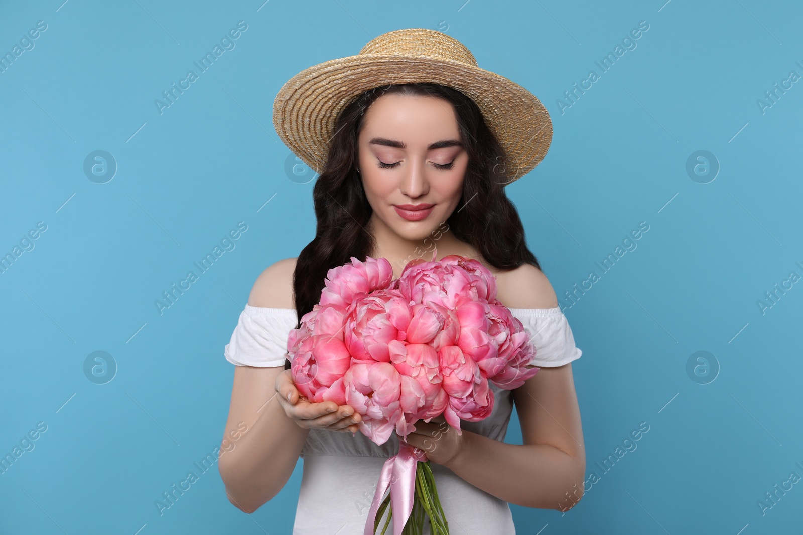 Photo of Beautiful young woman in straw hat with bouquet of pink peonies against light blue background