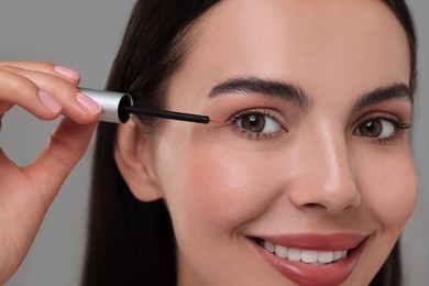 Photo of Beautiful woman applying serum onto her eyelashes on grey background, closeup. Cosmetic product