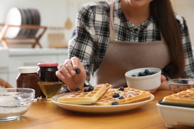 Photo of Woman decorating delicious Belgian waffles with blueberries at wooden table in kitchen, closeup