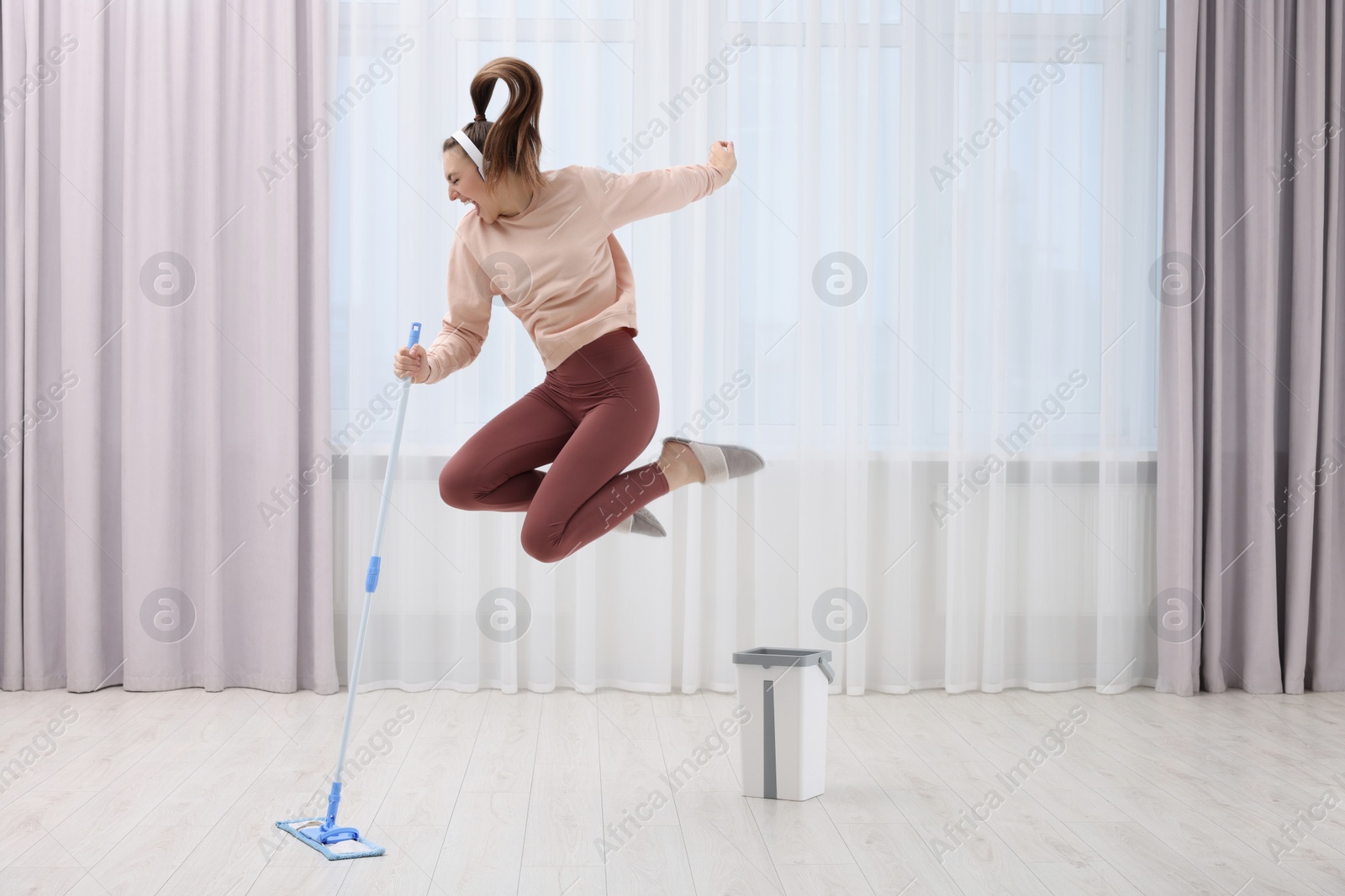 Photo of Enjoying cleaning. Woman in headphones jumping with mop indoors