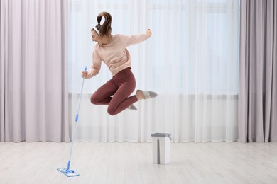 Photo of Enjoying cleaning. Woman in headphones jumping with mop indoors