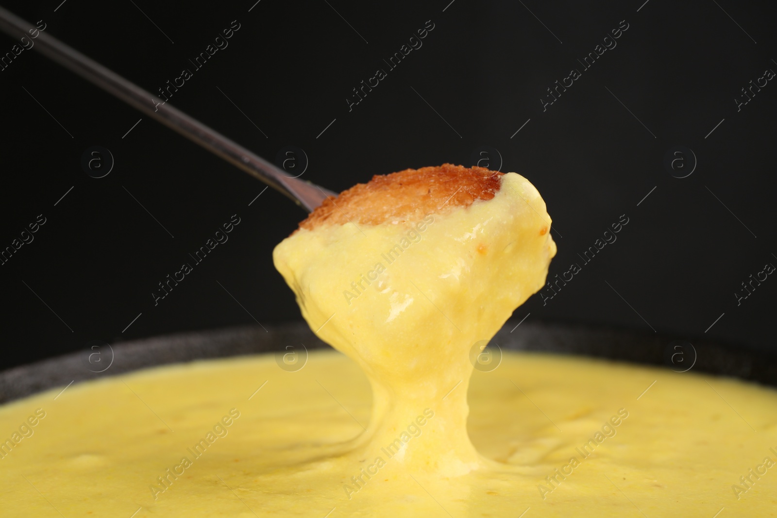 Photo of Dipping piece of bread into fondue pot with tasty melted cheese against dark gray background, closeup