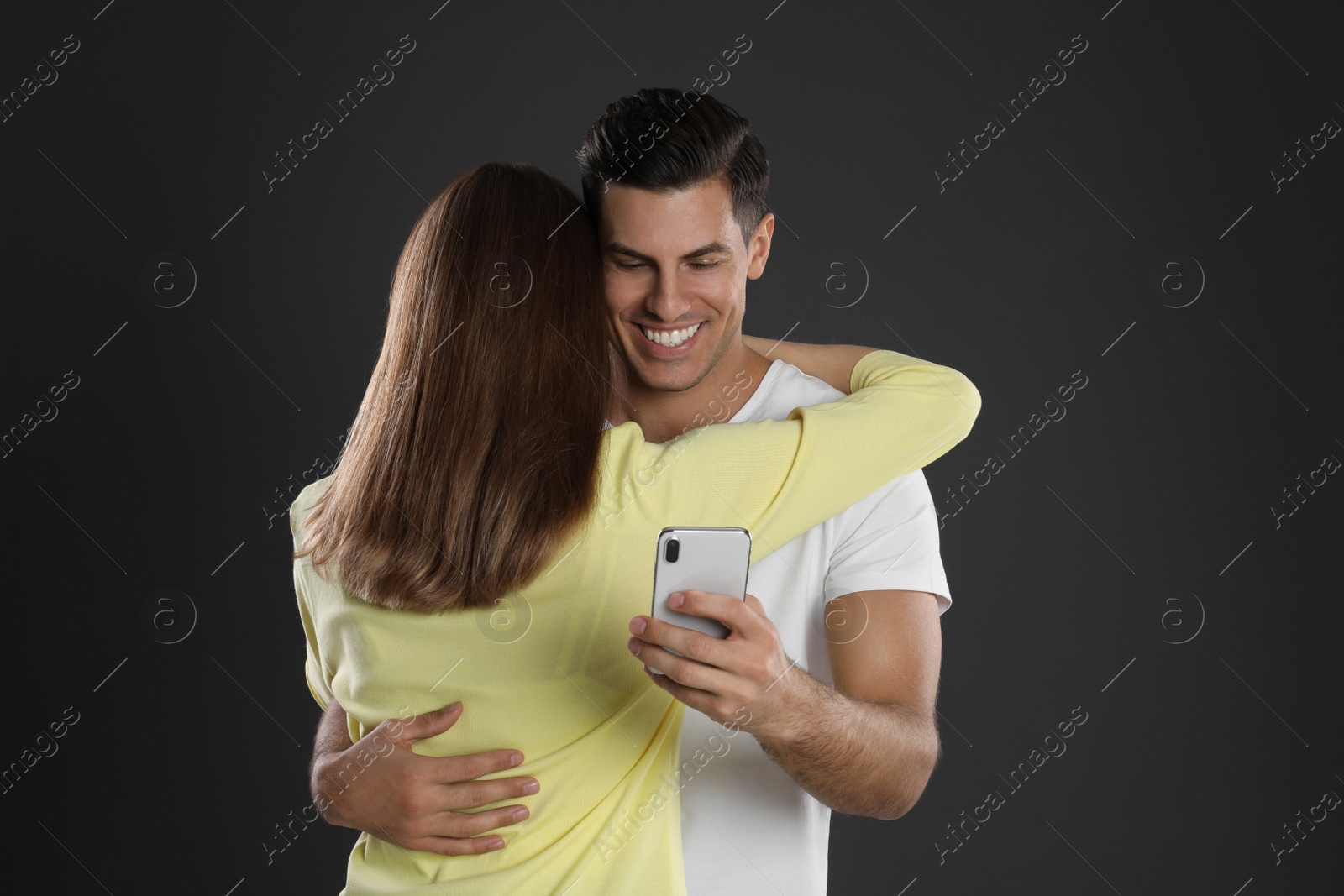 Photo of Man interested in smartphone while hugging his girlfriend on black background. Relationship problems