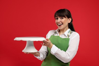 Happy professional confectioner in apron holding empty cake stand on red background