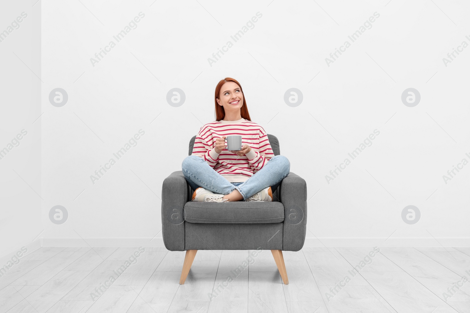 Photo of Happy young woman with cup of drink sitting in armchair near white wall indoors