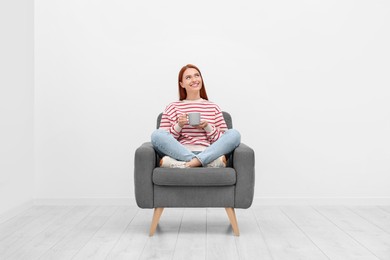 Photo of Happy young woman with cup of drink sitting in armchair near white wall indoors