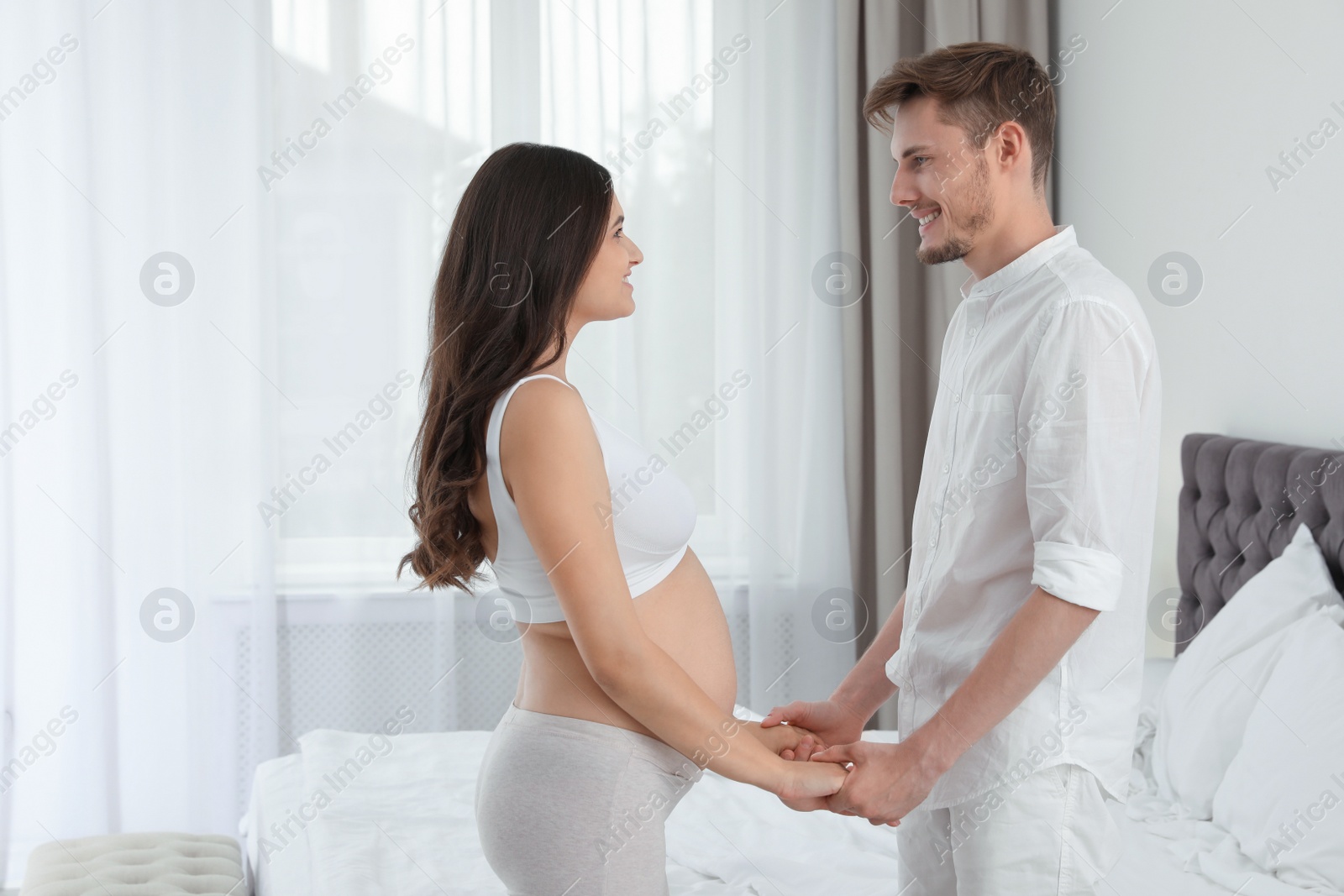 Photo of Young husband and his pregnant wife holding hands in bedroom