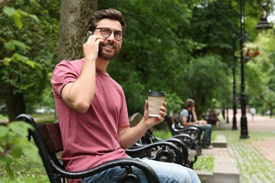 Photo of Handsome man with cup of coffee talking on smartphone in park