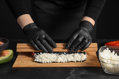 Photo of Chef in gloves wrapping sushi roll at dark textured table, closeup