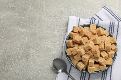 Photo of Brown sugar cubes in bowl and spoon on light grey table, flat lay. Space for text
