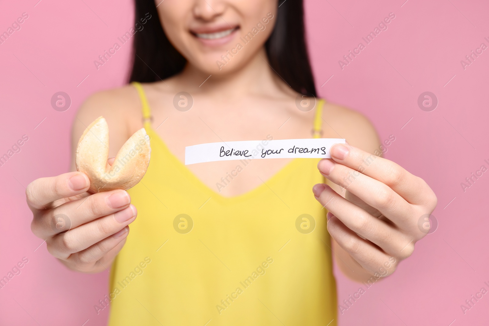 Photo of Young woman holding tasty fortune cookie with prediction Believe Your Dreams on pink background, closeup