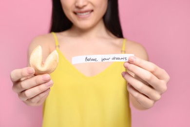 Young woman holding tasty fortune cookie with prediction Believe Your Dreams on pink background, closeup