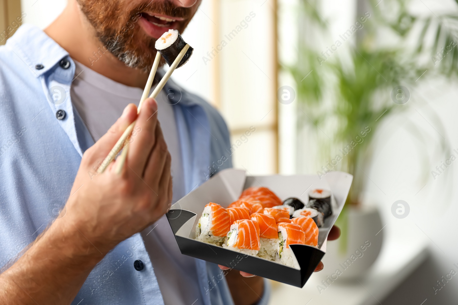 Photo of Handsome man eating sushi rolls with chopsticks indoors, closeup