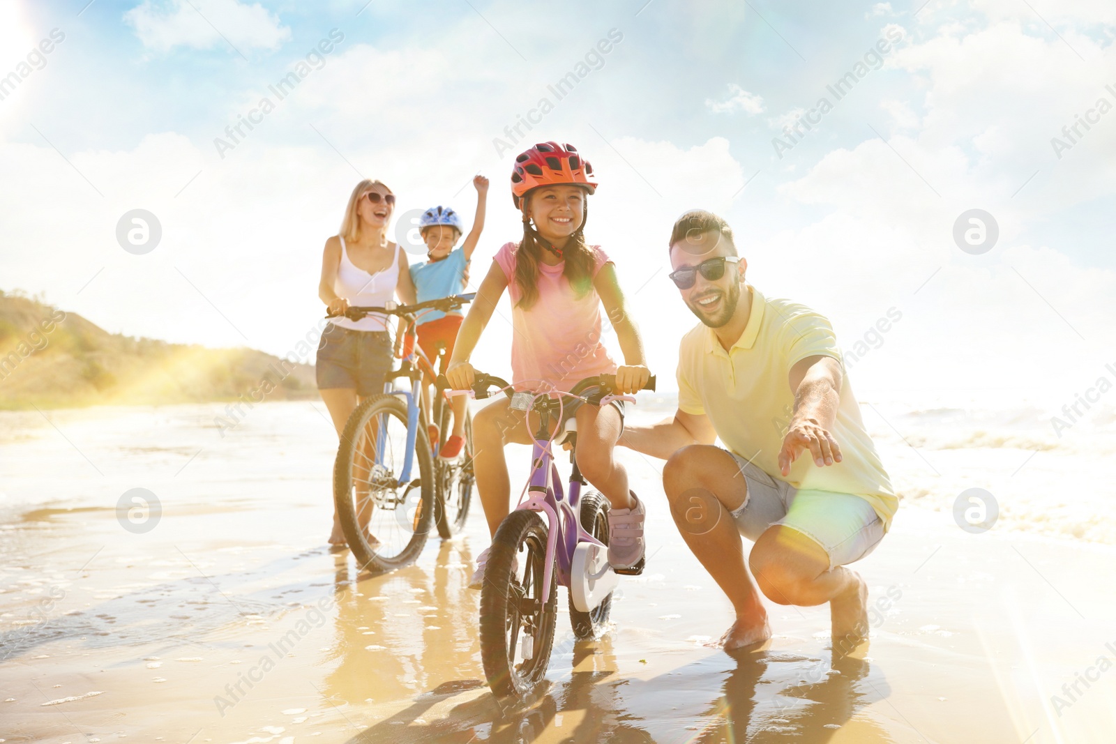 Image of Happy parents teaching children to ride bicycles near sea on sunny day