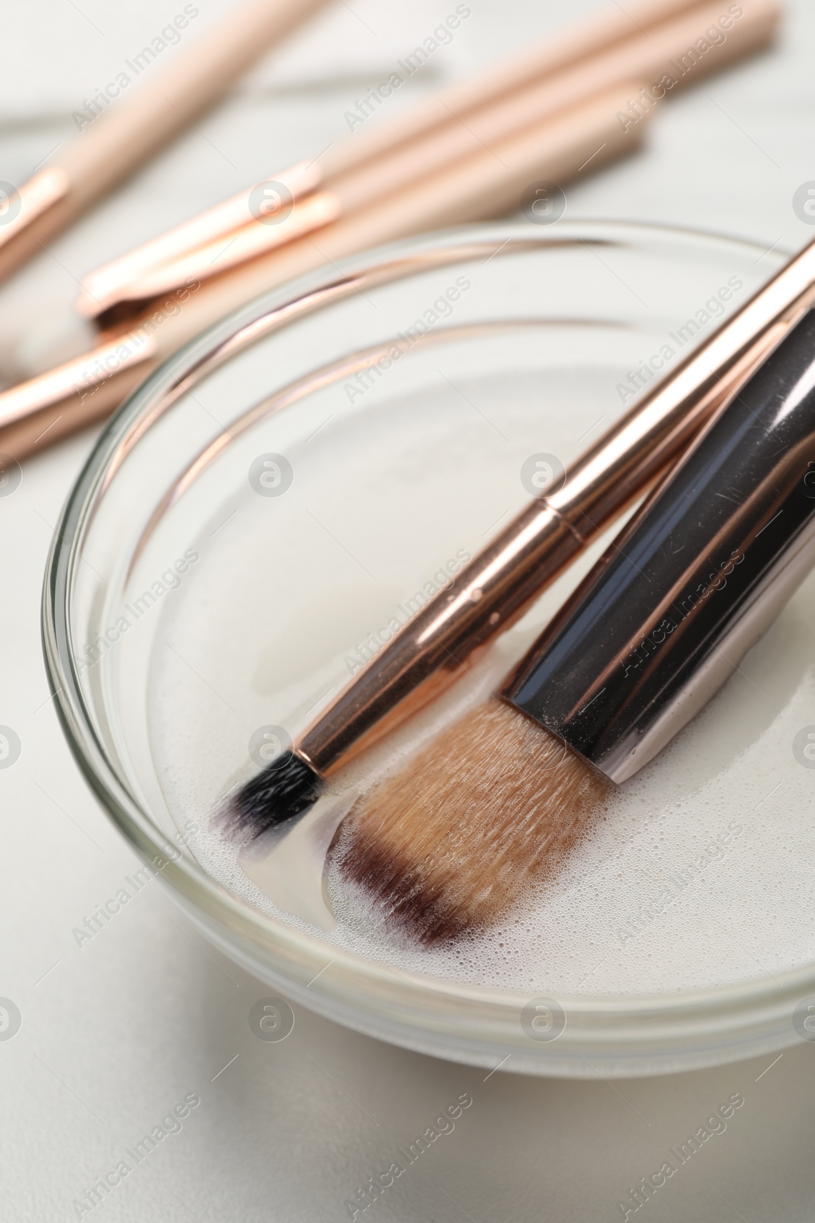 Photo of Clean makeup brushes in bowl with cleanser on white table, closeup