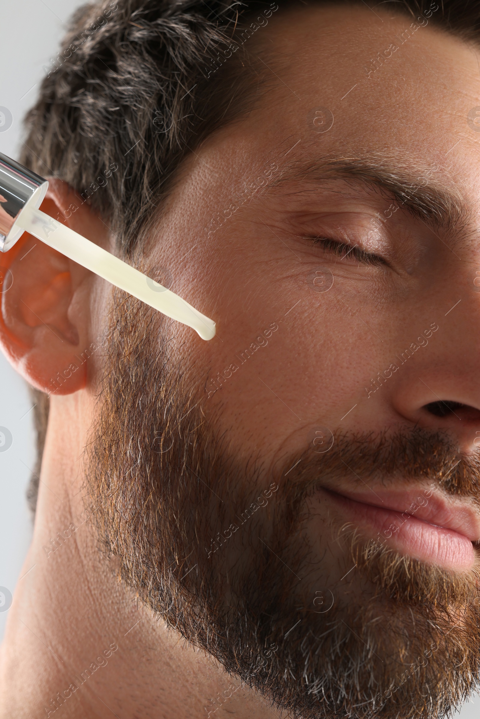 Photo of Handsome man applying cosmetic serum onto his face on light grey background, closeup