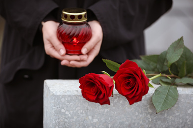 Woman holding candle near light grey tombstone with red roses outdoors, closeup. Funeral ceremony