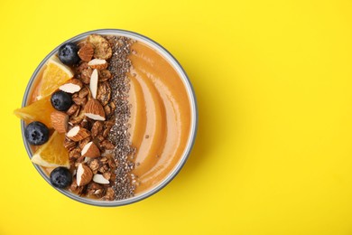 Bowl of delicious fruit smoothie with fresh orange slices, blueberries and granola on yellow background, top view. Space for text