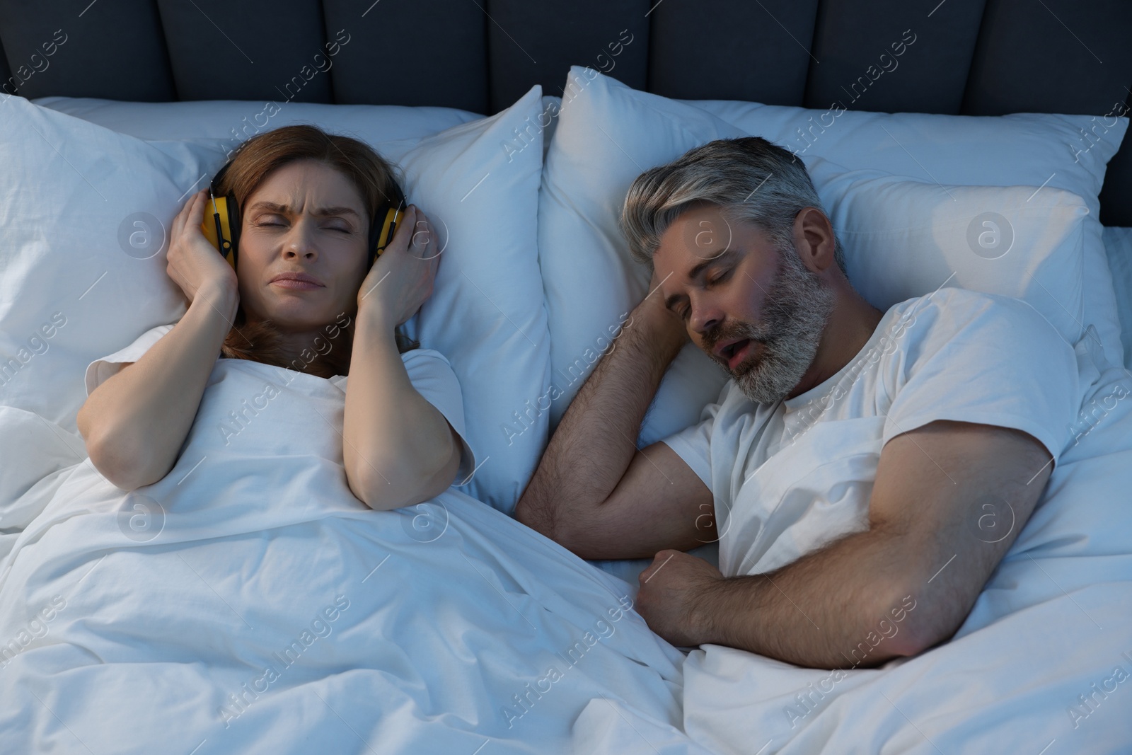 Photo of Irritated woman with headphones lying near her snoring husband in bed at home