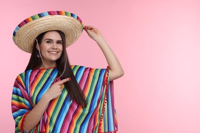 Photo of Young woman in Mexican sombrero hat and poncho pointing at something on pink background. Space for text